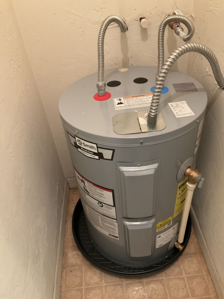 New electric water heater set on a black drain pan in a closet with flex connectors and a drain line