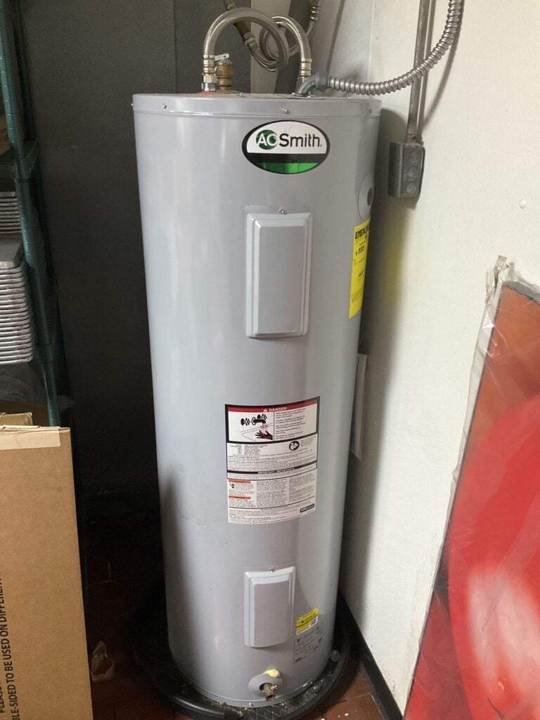 New tall electric water heater installed in a utility room with flexible conduit and water connections