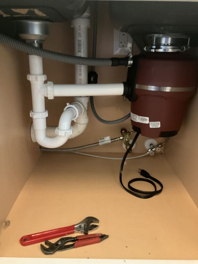 New garbage disposal mounted under a kitchen sink with PVC trap piping and a dishwasher hose