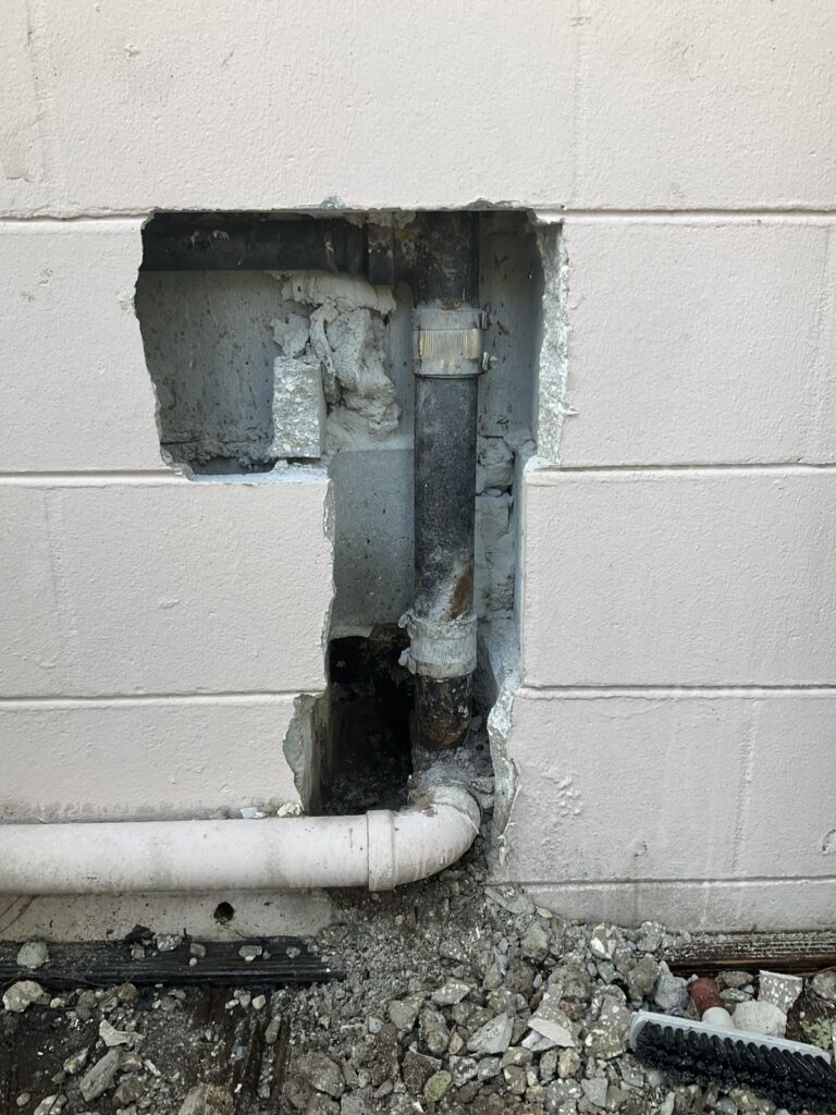Block wall opened at grade exposing an old galvanized riser and a white PVC line below