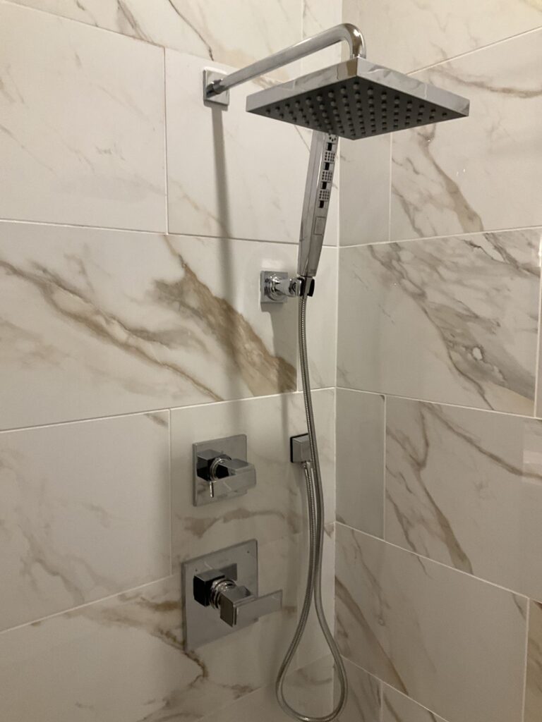 Chrome square rain shower head with a hand shower and two square trim valves on a marble tiled wall