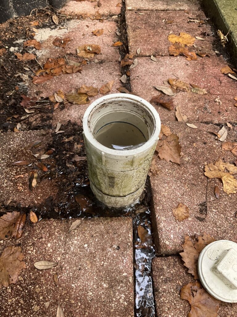 Water backing up out of an open sewer cleanout onto a brick walkway with the cap beside it