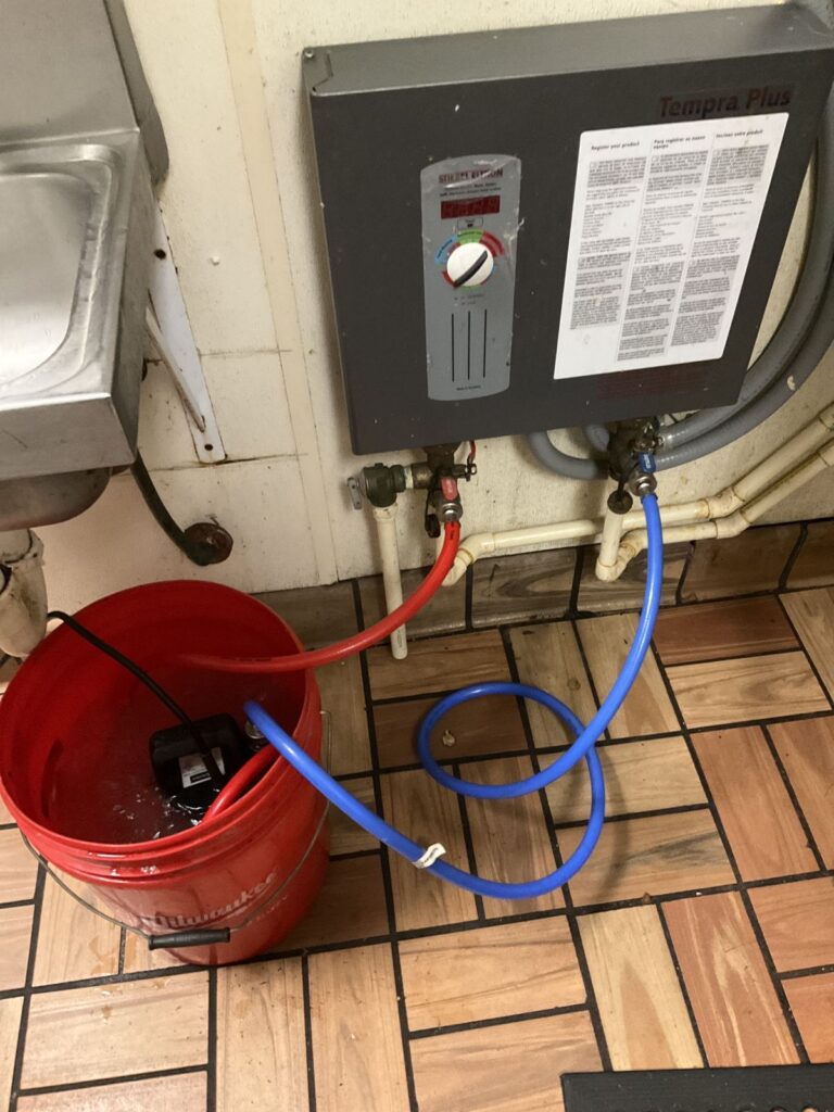 Electric tankless water heater being flushed with a pump, bucket and hoses on the isolation valves