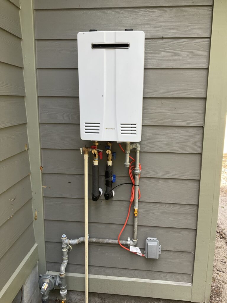 Exterior tankless water heater on gray siding with a gas line, isolation valves and an electrical box