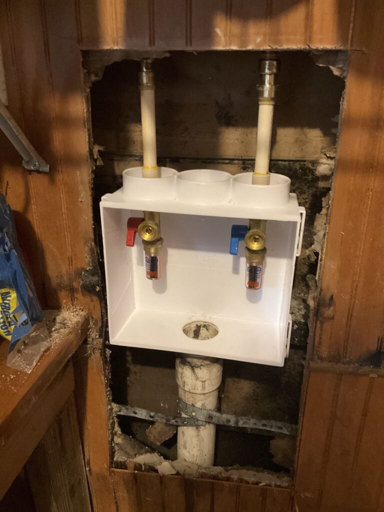 New white washing machine outlet box with hot and cold valves set in a wall opening above a drain