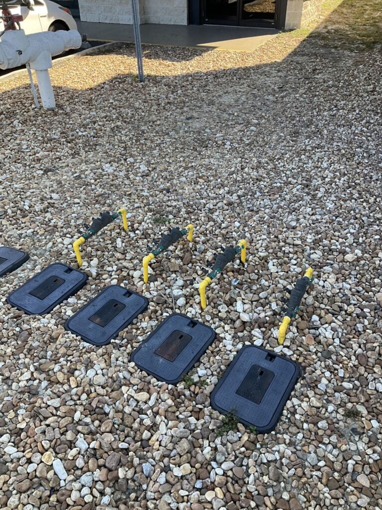 Row of yellow handled shutoff valves and valve boxes set in a gravel bed outside a building